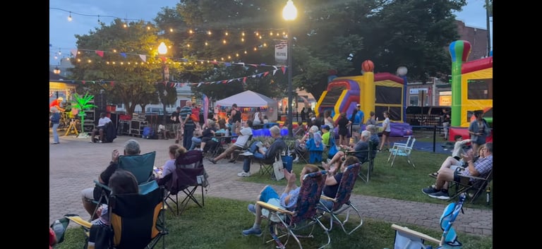 Outdoor evening event with families gathered on lawn, colorful inflatable structures, string lights overhead, and vendor tents