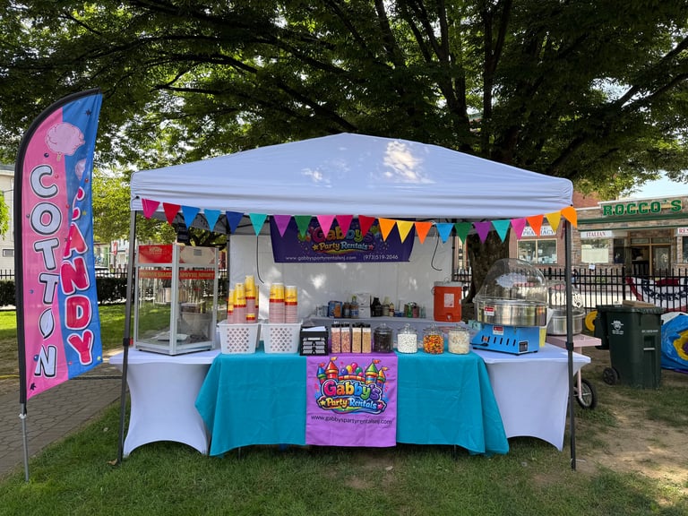 Outdoor market vendor booth with white canopy, colorful bunting, and display of cotton candy supplies and products on turquoise tablecloth