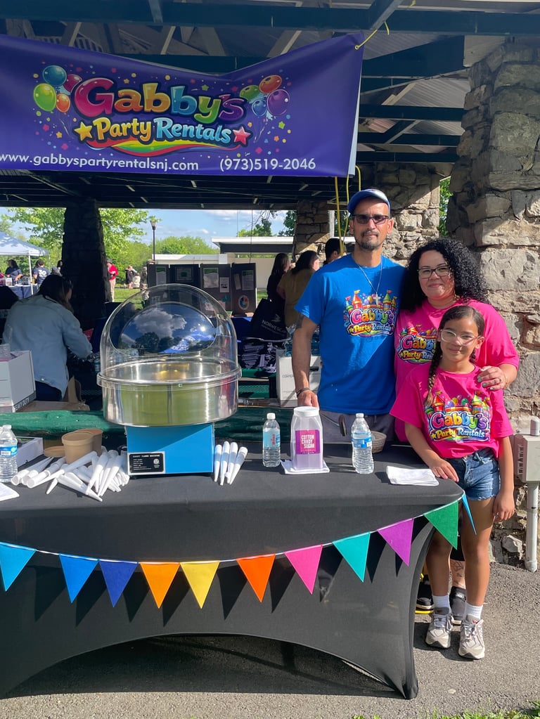 Gabbys Party Rentals booth with family staff at outdoor event, featuring popcorn machine and colorful banner