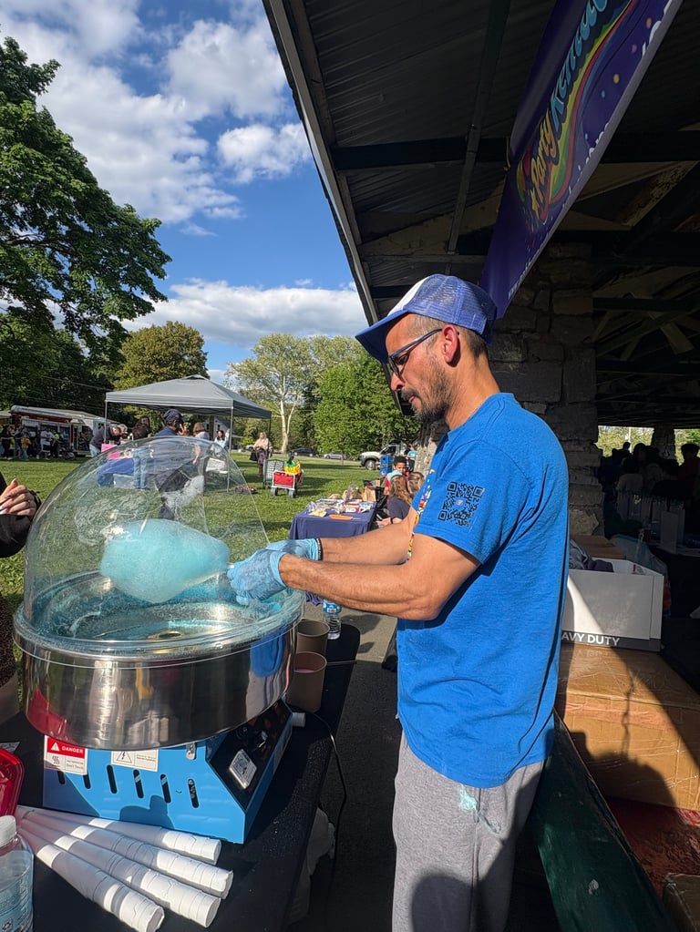 Man in blue shirt and cap operating a cotton candy machine at an outdoor event under a tent.