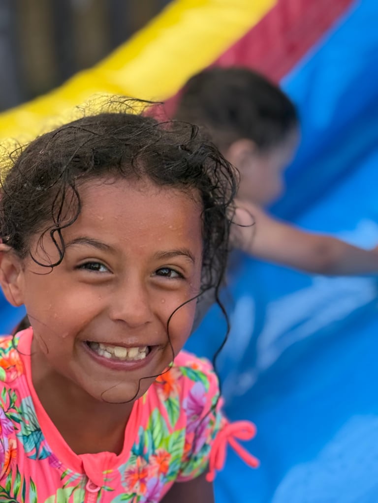 A smiling child in a bright pink colorful shirt enjoys a playground with blue and yellow equipment in the background