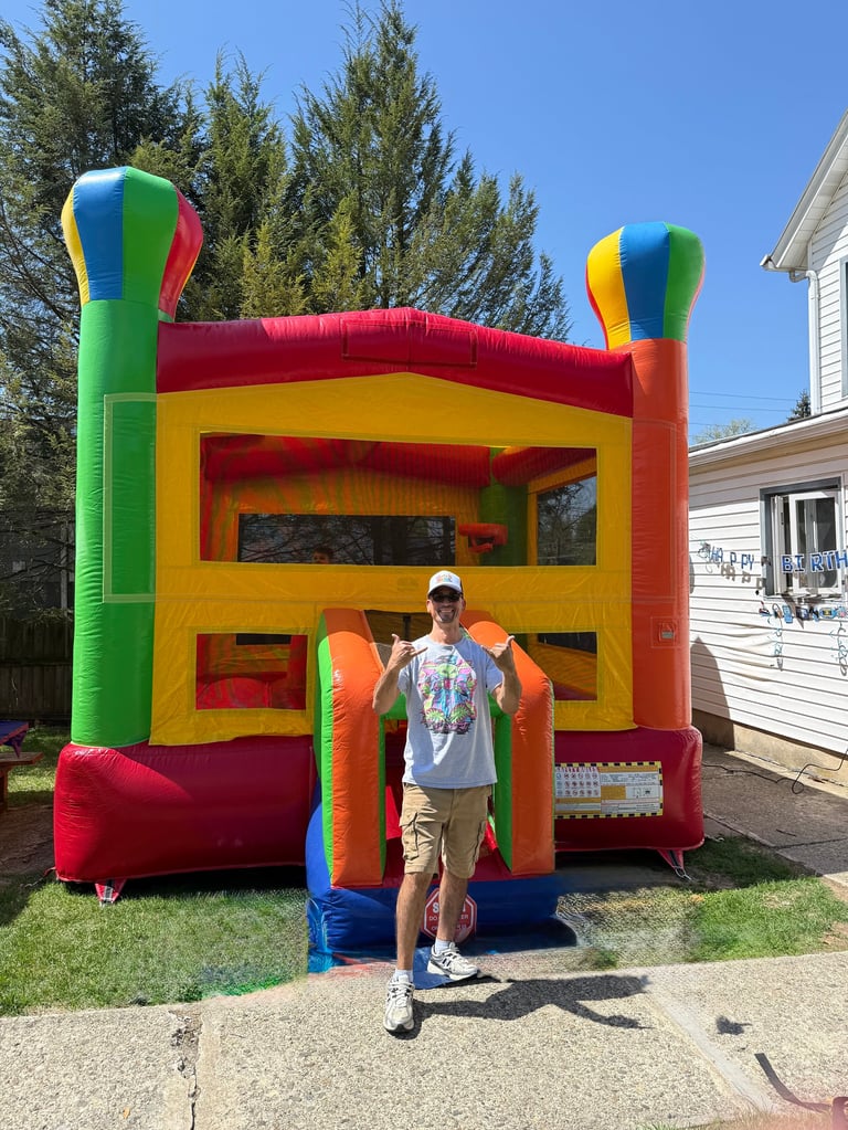 Man standing in front of a colorful inflatable bounce house with red, yellow, orange, and green sections in a residential driveway