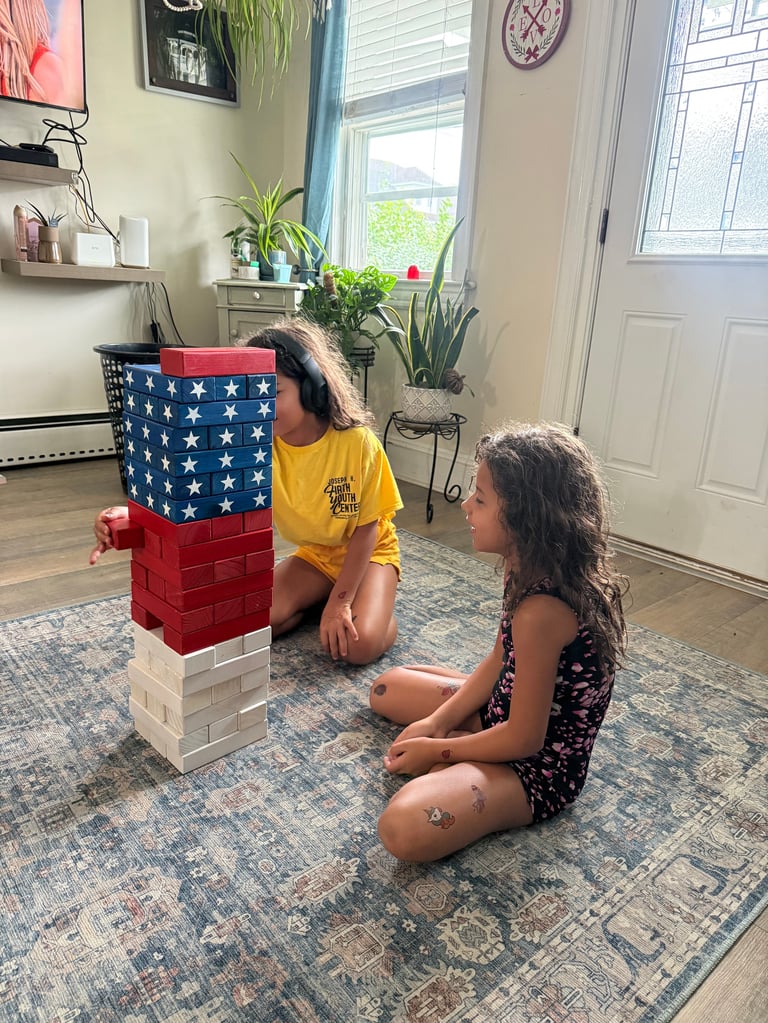 Two children playing with a patriotic stacking block tower in a bright living room with plants and windows