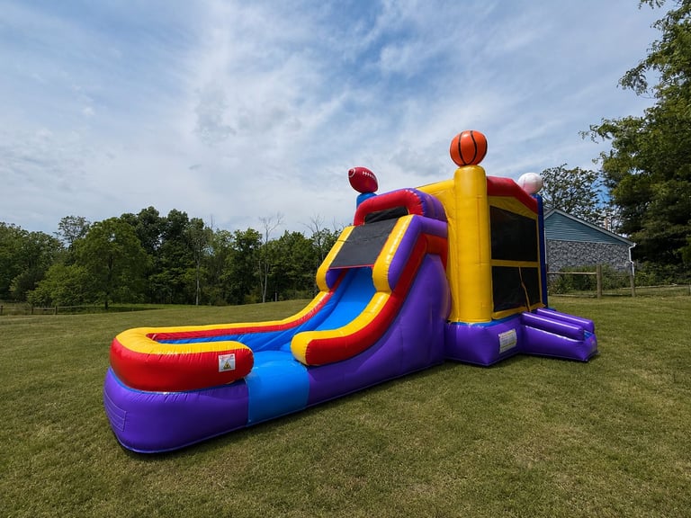 Colorful inflatable bounce house slide with purple, yellow, blue, and red sections on a grassy field under a sunny sky